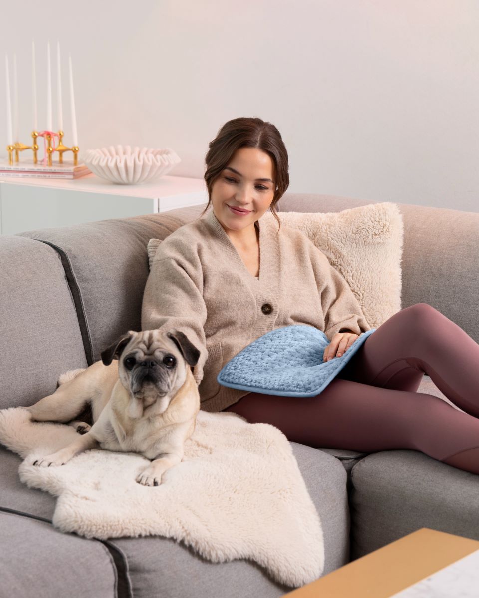 Woman sitting on the sofa with her dog while using a medisana heated pad on her lap.